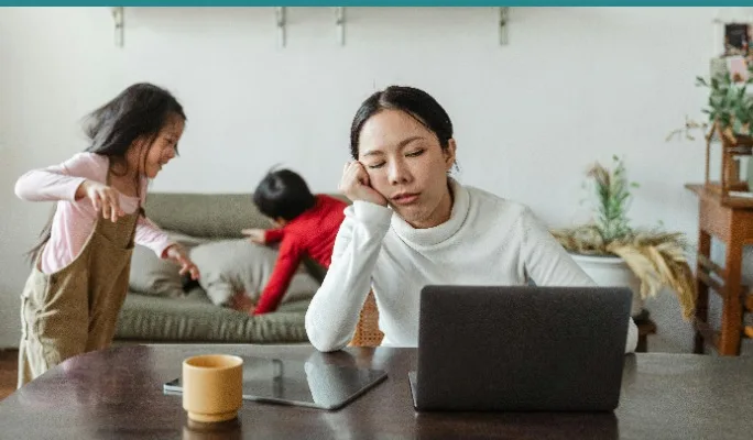 Tired mom at laptop while kids play on couch behind her