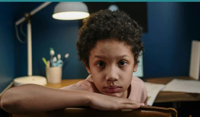 Girl sitting quietly at kitchen table, looking withdrawn while parent works nearby